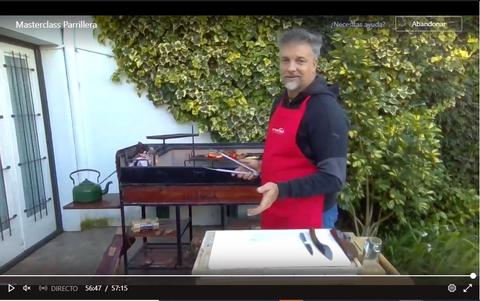 Un hombre sonriente con un delantal rojo se encuentra junto a un asador en un ambiente al aire libre.