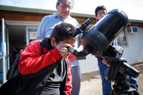 Un niño observa a través de un telescopio mientras un adulto y otro niño lo acompañan en un día soleado.