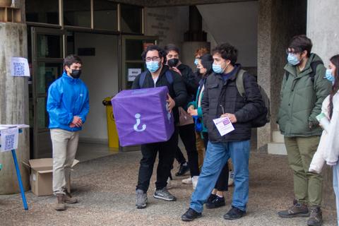 Grupo de estudiantes en una actividad al aire libre, con mascarillas y llevando una caja morada.