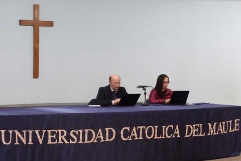 Dos personas están sentadas frente a computadoras portátiles en una mesa con un mantel que tiene el nombre de la universidad.