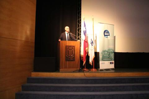 Un hombre habla desde un podio en un escenario con banderas nacionales y un cartel de evento.