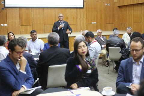 Una reunión en la que un hombre está hablando frente a un grupo de personas sentadas en mesas.
