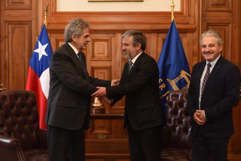 Tres hombres se encuentran en una reunión formal, con banderas de Chile y decoraciones institucionales visibles.