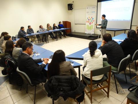 Un grupo de personas asiste a una presentación en un aula con mesas dispuestas en forma de U.