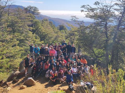 Un grupo grande de personas posando en un paisaje montañoso con árboles y un cielo despejado.