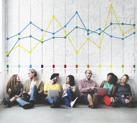 Un grupo diverso de personas sentadas en el suelo, trabajando en laptops y sonriendo, con un gráfico de líneas colorido en la pared detrás de ellos.