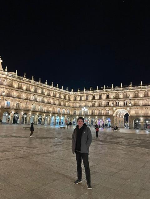 Una persona de pie en una plaza iluminada por la noche con edificios históricos de fondo.