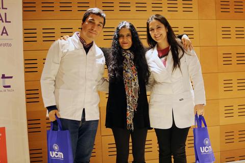 Tres personas posan juntas en un evento, dos de ellas con batas blancas y una con un abrigo negro.