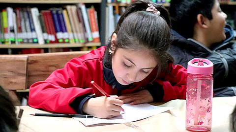 Una niña concentrada escribiendo en una mesa, rodeada de libros en una biblioteca.