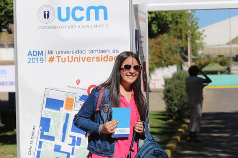 Una estudiante sonriente sostiene una libreta y se encuentra frente a un cartel de la Universidad Católica del Maule.