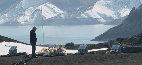 Una persona está de pie junto a tumbas adornadas con flores, con un paisaje montañoso cubierto de nieve y un lago al fondo.