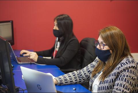 Dos mujeres trabajando en computadores mientras usan mascarillas en una oficina.