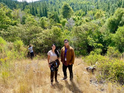 Dos personas posan en un paisaje natural rodeado de vegetación y árboles.