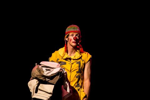 Un payaso con un gorro colorido y maquillaje de payaso está de pie en un escenario oscuro con una mochila en la mano.