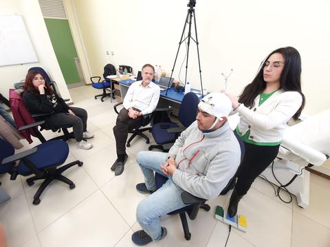 Un grupo de personas en un laboratorio observan a una mujer que está colocando un dispositivo en la cabeza de un hombre.