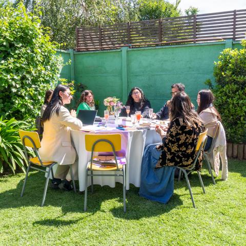 Un grupo de personas sentadas alrededor de una mesa en un entorno al aire libre, rodeadas de plantas y flores.