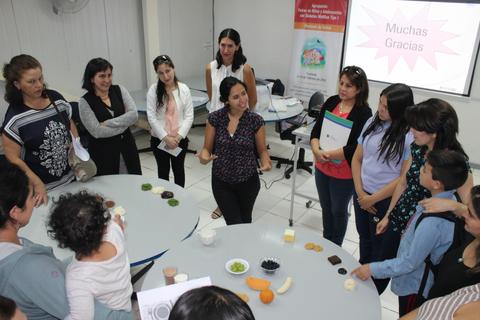 Un grupo de personas en un taller o presentación, observando a una mujer que explica mientras se aprecian varios ingredientes en una mesa.