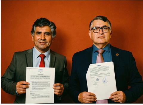 Dos hombres posan con documentos importantes en un fondo colorido.