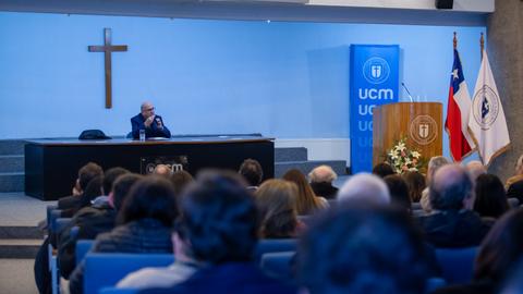 Una conferencia en la UCM con una cruz de fondo y un público atento.