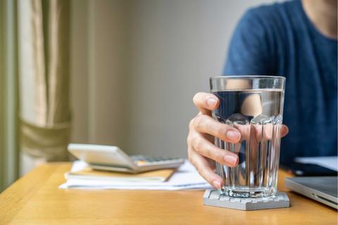 Una persona sostiene un vaso de agua en una mesa con documentos y una calculadora al fondo.