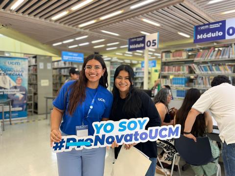 Dos estudiantes sonrientes sosteniendo un cartel en una biblioteca universitaria.
