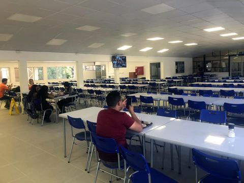 Una cafetería con mesas y sillas vacías y algunas personas sentadas.