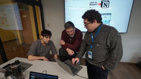 Tres personas colaboran frente a una computadora en una sala de trabajo.