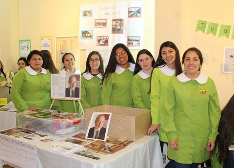 Un grupo de siete mujeres vestidas con batas verdes sonríen mientras están junto a una mesa con diferentes materiales y fotografías.