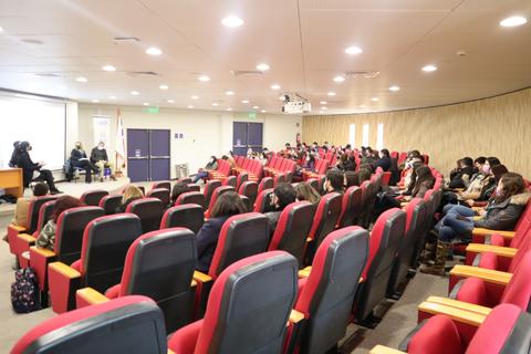 Una sala de conferencias con asistentes sentados en asientos rojos y un panel de discusión al frente.