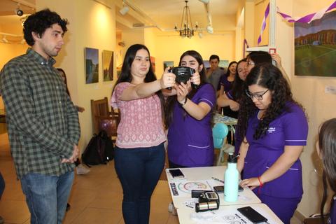 Un grupo de jóvenes interactúa en un evento, mientras una persona toma una foto con una cámara.