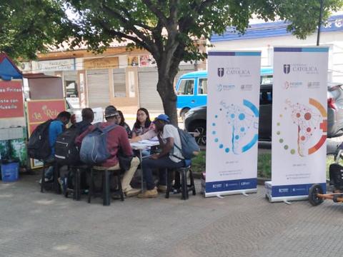 Un grupo de jóvenes se reúne en una mesa al aire libre, rodeados de banners informativos y un árbol, en un entorno urbano.