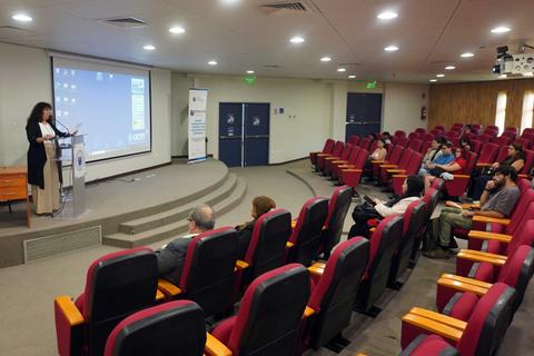 Una expositora presenta su tema frente a un público en un auditorio.