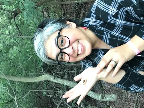 Una mujer sonriente sostiene un insecto en su mano mientras está rodeada de árboles.