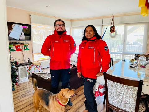 Dos personas con chaquetas rojas posan en una sala con un perro y decoraciones navideñas.