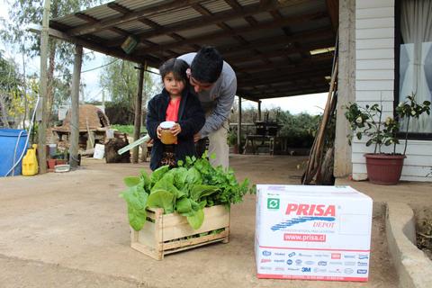 Una niña sostiene una taza mientras un adulto se inclina hacia ella en un espacio al aire libre con plantas y una caja de la empresa PRISA.