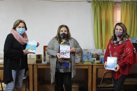 Tres mujeres sosteniendo libros en un ambiente de aula con libros al fondo.