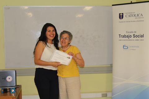 Una joven y una mujer de edad avanzada posan sonrientes sosteniendo un certificado en un aula.