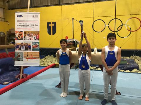 Tres niños en un gimnasio están posando con medallas y un trofeo, rodeados de equipo de gimnasia.