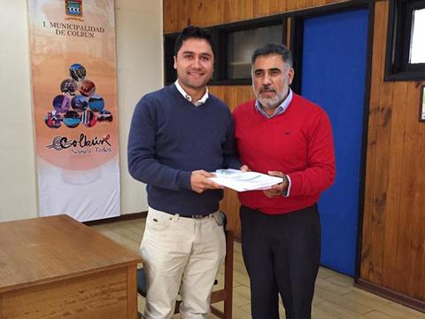 Dos hombres se están saludando y uno de ellos sostiene unos documentos en un ambiente formal.