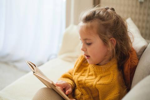 Una niñita está leyendo un libro cómodamente en el sofá.