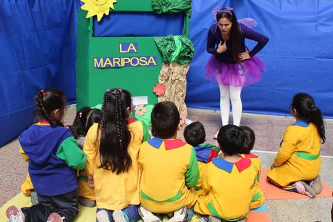 Una profesora cuenta una historia a un grupo de niños sentados en el suelo frente a un escenario decorado.