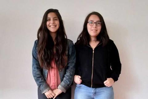 Dos mujeres posan juntas frente a una pared blanca, sonriendo y vestidas de manera casual.