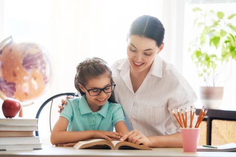 Una madre sonriente ayuda a su hija a leer un libro en un ambiente acogedor.
