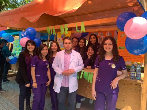 Grupo de personas en un evento, todas vistiendo uniformes morados y sonriendo frente a un stand decorado con globos azules y rosas.