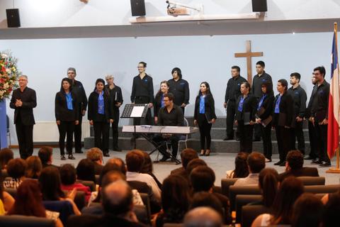 Un coro está cantando en una ceremonia en una iglesia, con un piano y un crucifijo al fondo.