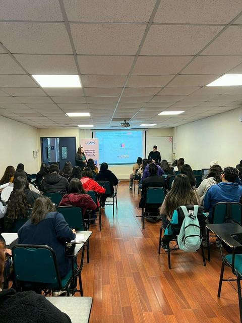 Una sala de clases con estudiantes sentados en escritorios y un instructor frente a una proyección.