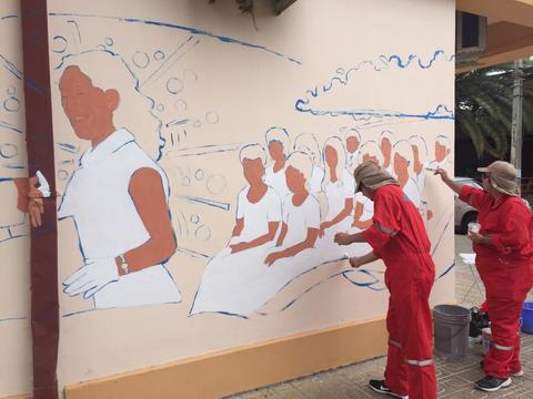 Un mural en proceso de pintura, con varias figuras humanas representadas y dos personas trabajando en la obra.