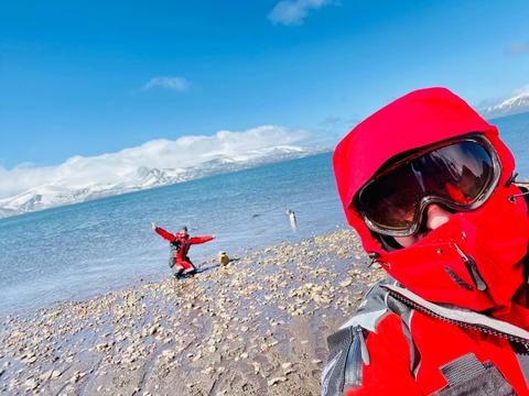 Dos personas en una playa helada, una en primer plano con abrigo rojo y otra al fondo divirtiéndose.
