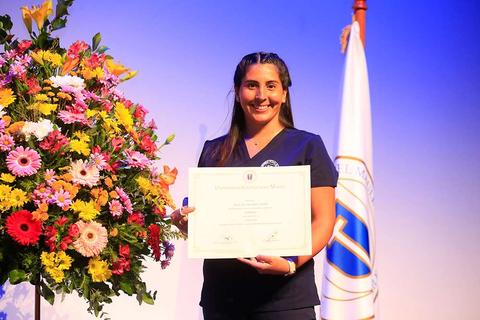 Una mujer sonriente sostiene un certificado en un escenario decorado con flores coloridas.