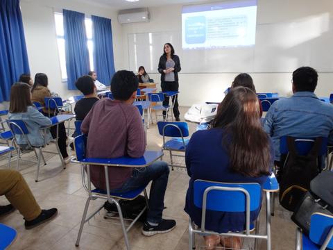 Una mujer está presentando frente a una clase llena de estudiantes sentados en escritorios.